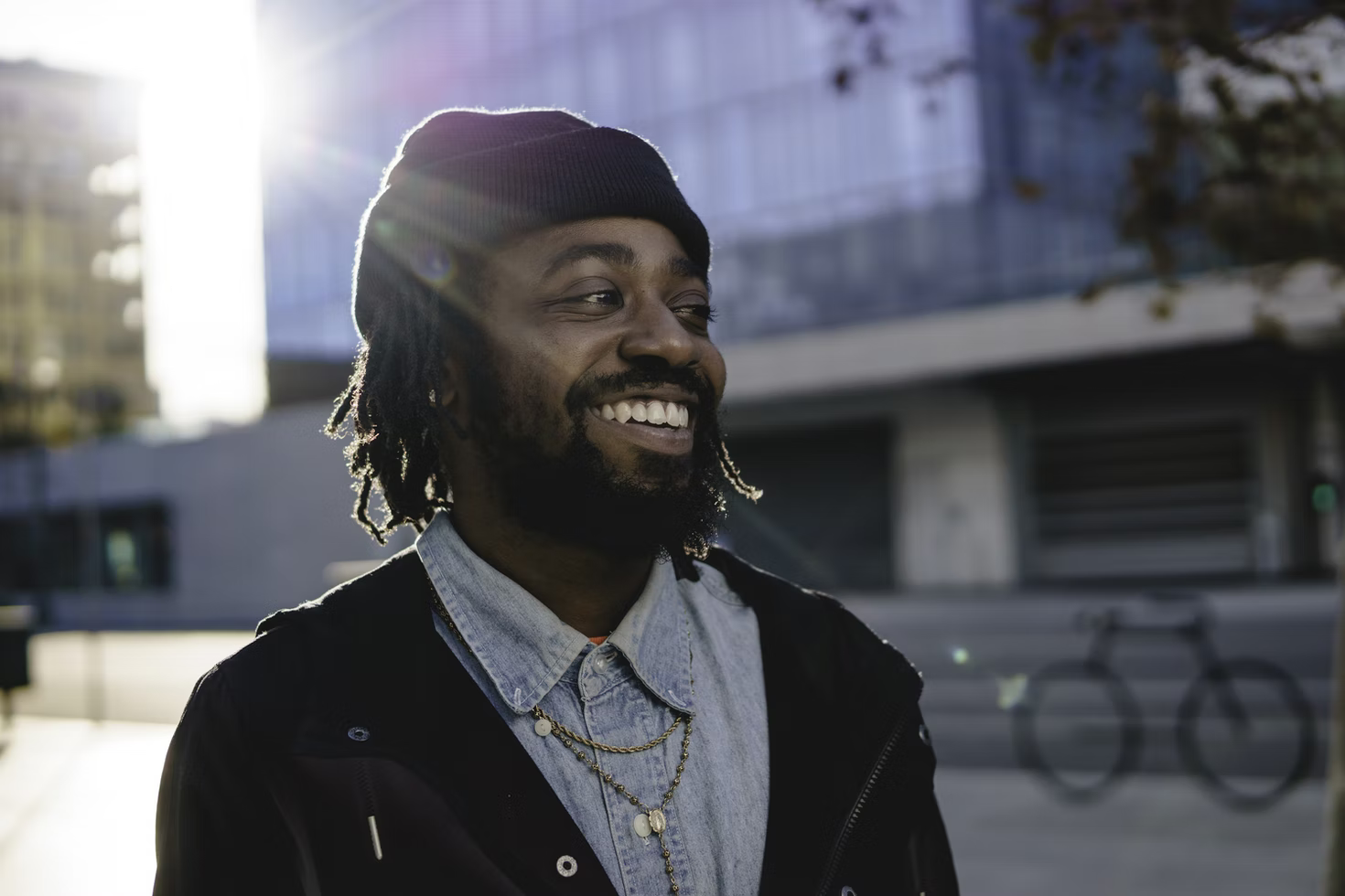A smiling man with dreadlocks and a cap standing outside.