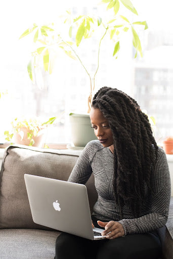 Woman with very long, thick locs using a laptop on a couch. 