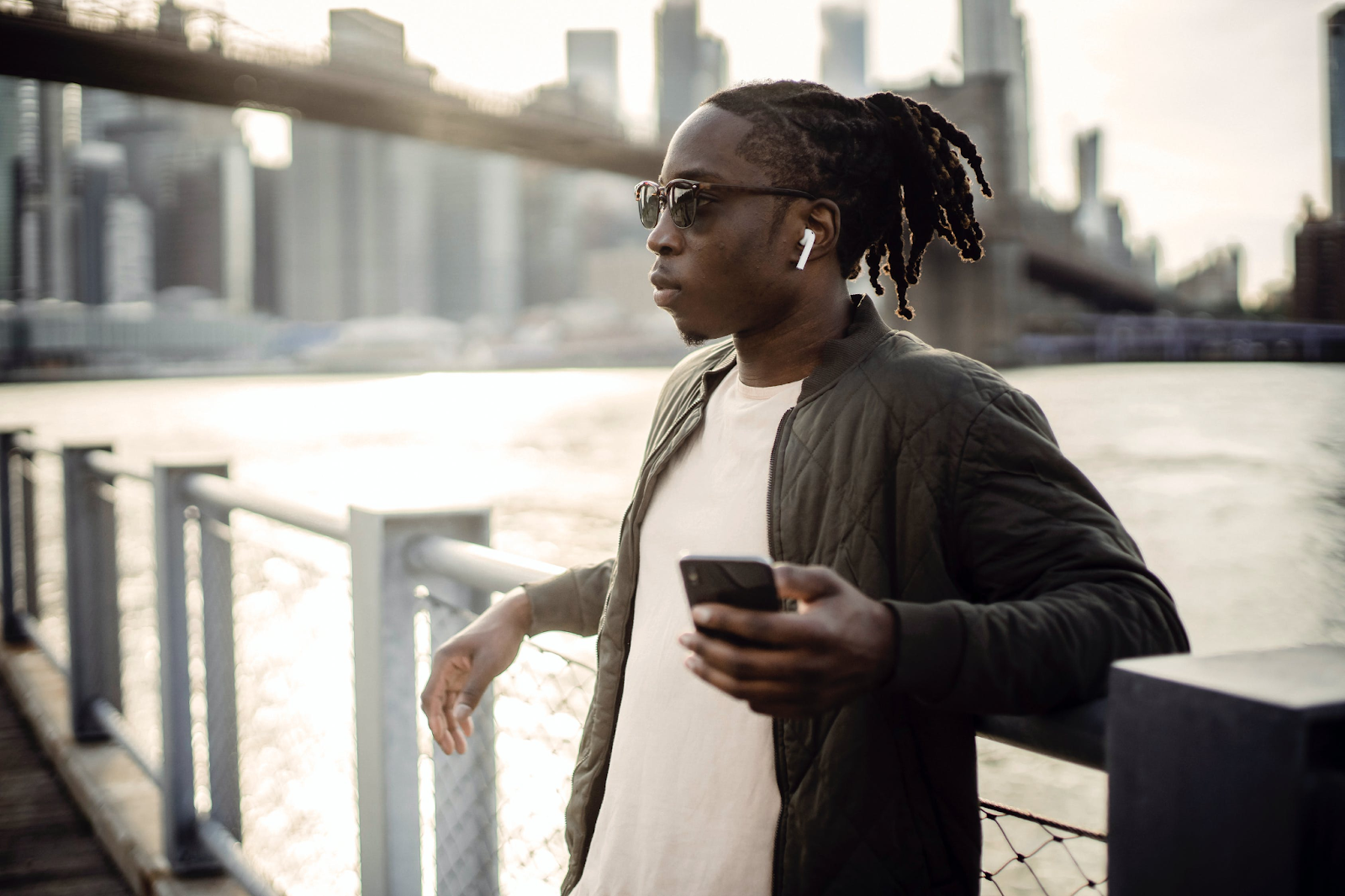Well dressed black man with a dreadlock ponytail and sunglasses relaxing by a bridge port.