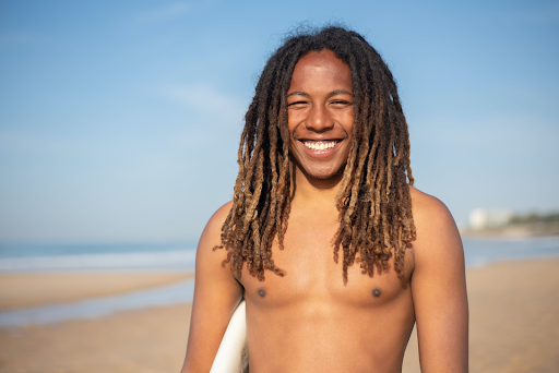 Smiling, shirtless man with long dreadlocks at the beach.